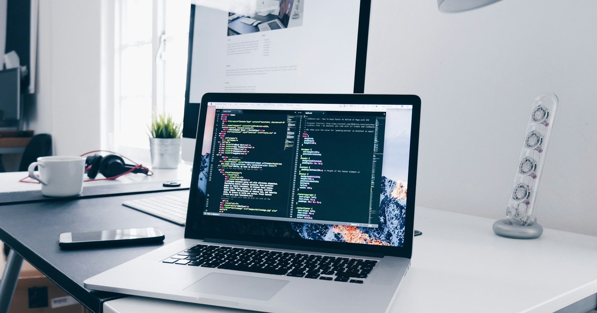 Laptop showing code on a white desk beside a monitor, phone, mug, headphones and a small plant